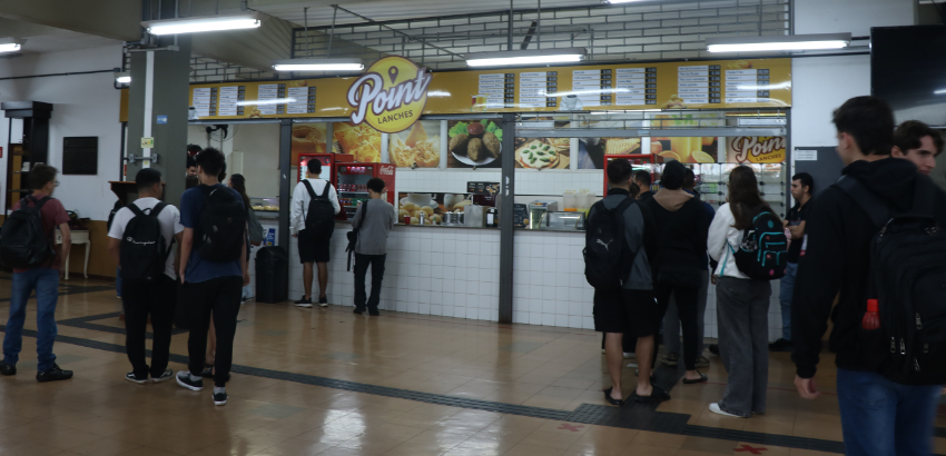 Imagem com fundo de uma lanchonete em um ambiente de circulação. Também encontra pessoas andando e comendo em torno do local. 