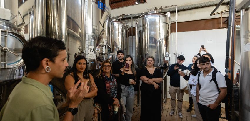 Grupo de pessoas participa de visita guiada em cervejaria, com tanques de inox ao fundo; guia explica processo enquanto visitantes observam e registram com celulares.