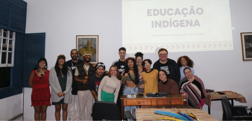 Grupo de estudantes e participantes indígenas posam juntos em frente a um projetor com a apresentação “Educação Indígena” durante evento acadêmico em uma sala com paredes brancas e janelas azuis.