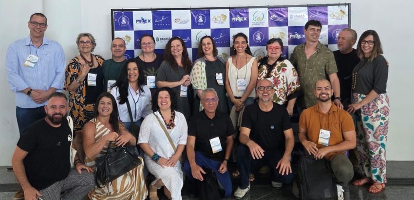 Grupo de profissionais posa sorridente em frente a painel institucional durante evento acadêmico ou cultural. A diversidade de expressões e estilos transmite um clima descontraído e colaborativo.