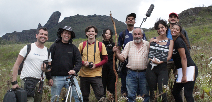 Grupo de nove pessoas posa sorridente, durante as filmagens. Um integrante segura um cajado, outro uma claquete, e há equipamentos de filmagem visíveis. Ao fundo, montanhas e céu encoberto. 