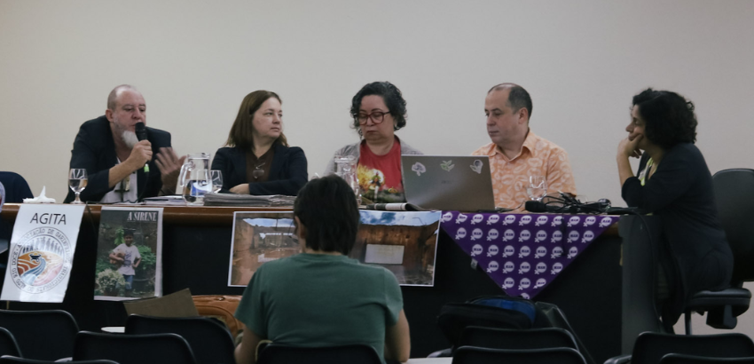 Mesa-redonda com cinco participantes em um auditório. Um deles fala ao microfone enquanto os demais acompanham. Há cartazes e fotos expostos na frente da mesa e uma pessoa do público aparece de costas
