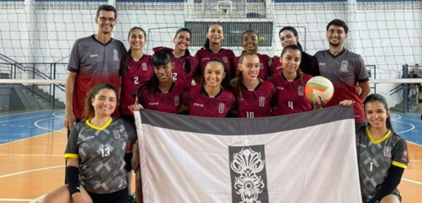 Time feminino de voleibol posa em quadra coberta, com jogadoras uniformizadas em vermelho e preto e dois membros da comissão técnica ao fundo. Duas atletas ajoelhadas na frente seguram uma bandeira branca com um símbolo central, enquanto outra segura uma 