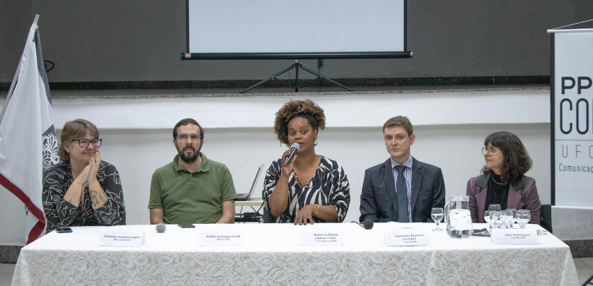 Cinco pessoas (três mulheres e dois homens) compõem uma mesa. Uma mulher ao centro fala ao microfone, enquanto os demais escutam.