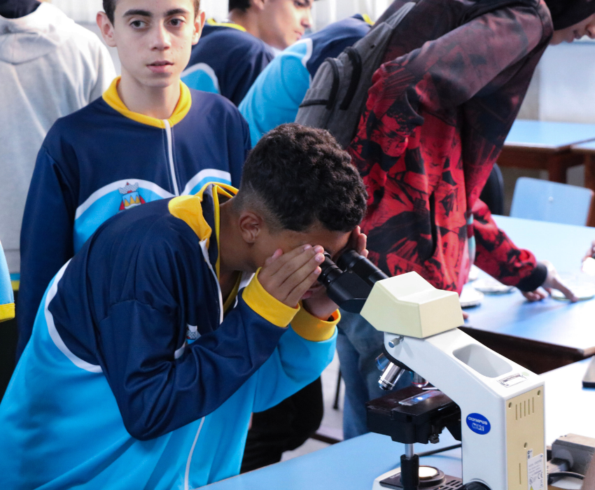 Imagem mostra aluno uniformizado na cor azul e detalhes em amarelho. Ele observa fungos ao microscópio, ao fundo é visível outros alunos na demais áreas da mostra.