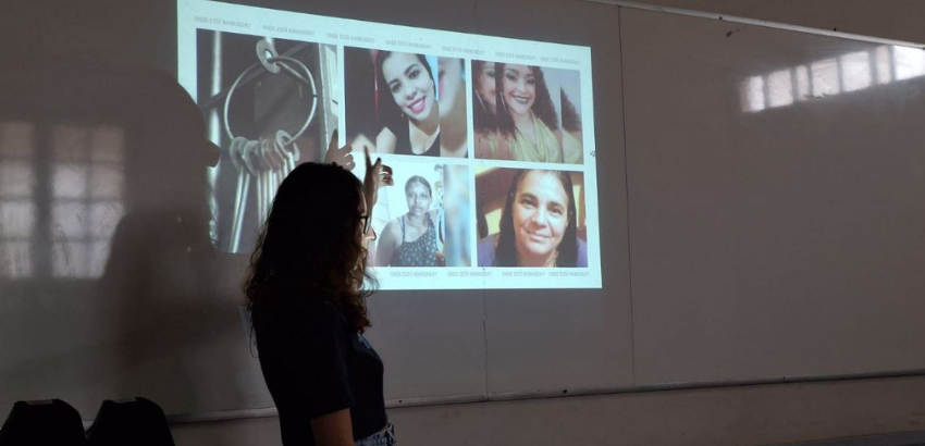 Imagem mostra integrante do Ariadnes. Ela ministra uma palestra formativa e aponta para uma imagem quatro mulheres de diferentes idades e um conjunto de chave. Ao fundo um quadro branco. 