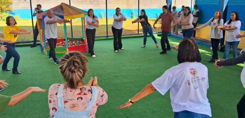Imagem mostra grupo participando de prática de atividade física. Dezessete pessoas compõem a roda. Observamos um fotógrafo ao fundo e um grupo de pessoas ao fundo conversando. 