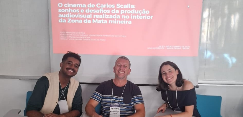 Três pessoas sorrindo sentadas em uma sala de aula, diante de uma tela com o texto “O cinema de Carlos Scalla: sonhos e desafios da produção audiovisual realizada no interior da Zona da Mata mineira”.
