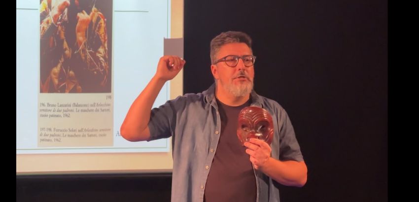 Homem de óculos, barba grisalha e camisa azul aberta sobre camiseta escura faz uma apresentação em um palco. Ele segura uma máscara marrom esculpida em uma das mãos e gesticula com a outra enquanto fala. Ao fundo, há uma tela de projeção ex