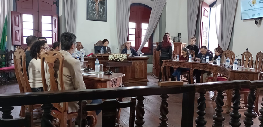 Reunião formal em uma sala, com pessoas sentadas e dispostas em formato de semicírculo. Ao fundo, há janelas grandes com cortinas, uma mesa com três pessoas e um telão exibindo informações.