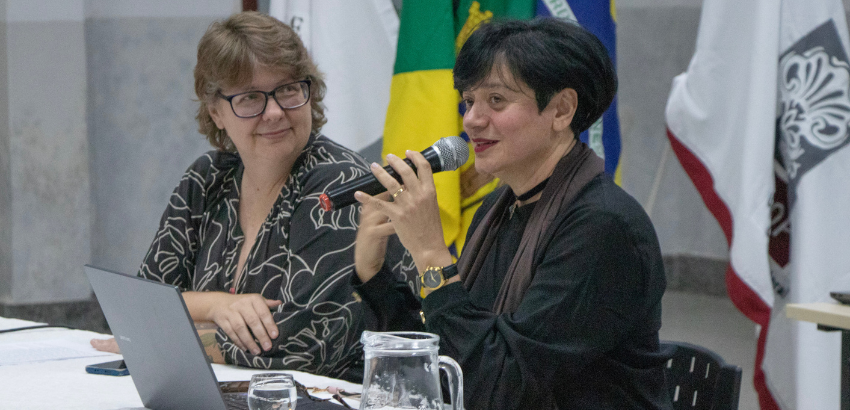 Duas mulheres sentadas à mesa durante uma conferência. Uma fala ao microfone, enquanto a outra observa sorrindo.