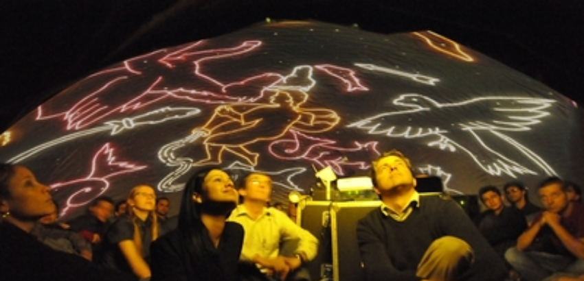 rupo de pessoas sentadas dentro de um planetário, olhando para uma projeção colorida no teto em forma de constelações estilizadas, incluindo figuras humanas e aves delineadas com luz