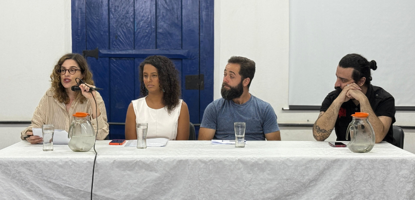 Mesa com quatro pessoas sentadas, duas mulheres e dois homens, participando de uma discussão. A mulher à esquerda fala ao microfone. Há jarros de vidro e copos sobre a mesa coberta com toalha branca. Uma porta azul ao fundo