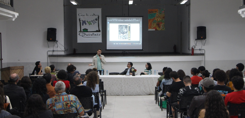 Imagem mostra o professo e jornalista, João Marcos em pé com microfone na mão, estudantes sentados e de consta e ao fundo banner do grupo quintais durante o primeiro simpósio nacional de jornalismo de quadrinhos, na UFOP, em Mariana. 