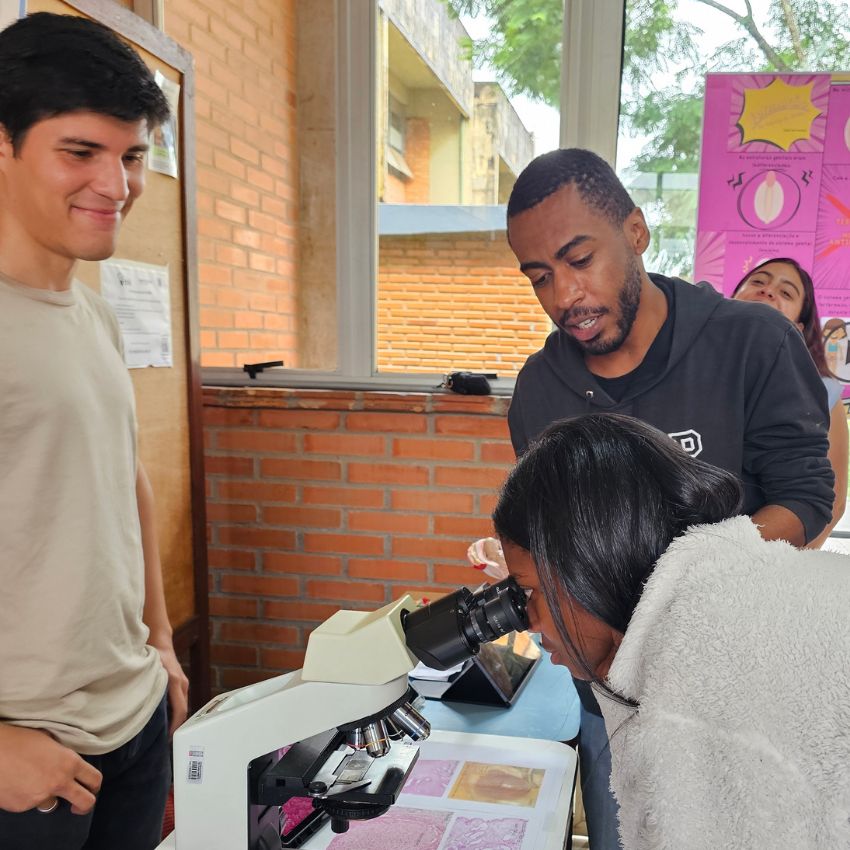 Estudante observa lâmina em microscópio enquanto dois estudantes de medician a acompanham, em sala com paredes de tijolo e janela ao fundo.