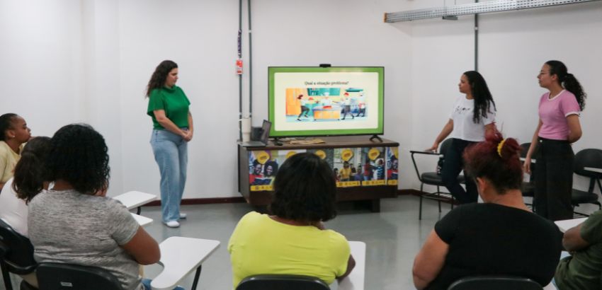 rês jovens apresentam em sala de aula para um grupo de pessoas sentadas em carteiras. No telão aparece a pergunta “Qual a situação problema?” com ilustração colorida. A atividade ocorre em ambiente de formação ou oficina educativa.
