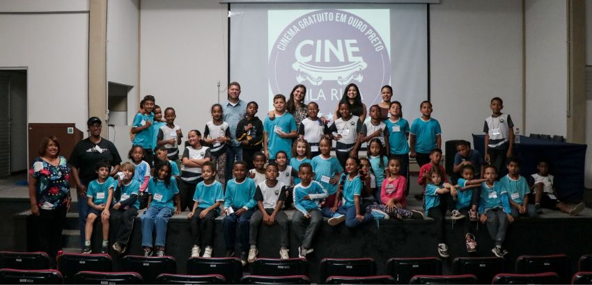 Grupo de crianças e adultos posa sorrindo em auditório diante de telão com o logotipo “Cine na Rua – Cinema gratuito em Ouro Preto”. As crianças vestem camisetas azuis e brancas, e estão acompanhadas por educadores e organizadores do evento.