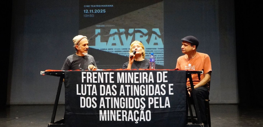 Três pessoas ministram uma mesa. Sob a mesa há uma faixa escrito "Frente Mineira de Luta das Atingida e Atingidos pela Mineiração"