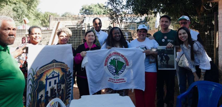 "Grupo de cerca de dez pessoas reunidas ao ar livre, sorrindo para a câmera. No centro, participantes seguram uma bandeira do Movimento dos Atingidos por Barragens (MAB). À esquerda, há um painel com a inscrição 'Comunidade Quilombola'. Ao fundo, árvores,