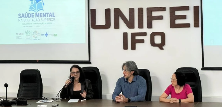 Mesa em auditório composta por duas mulheres e um homem ao centro. A mulher da esquerda está de roupas pretas e falando ao microfone, o homem é grisalho e veste azul e mulher da direita está vestida com camiseta rosa.