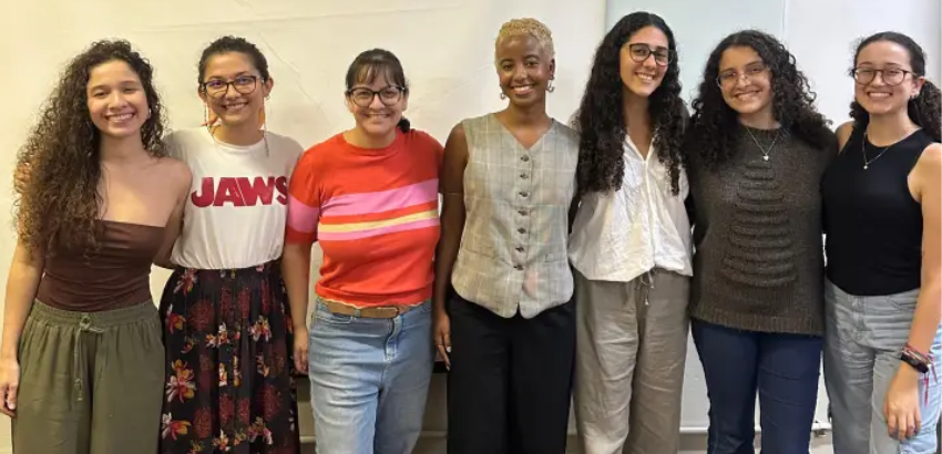 Grupo de sete mulheres lado a lado, sorrindo para a câmera, em ambiente interno de parede clara. 