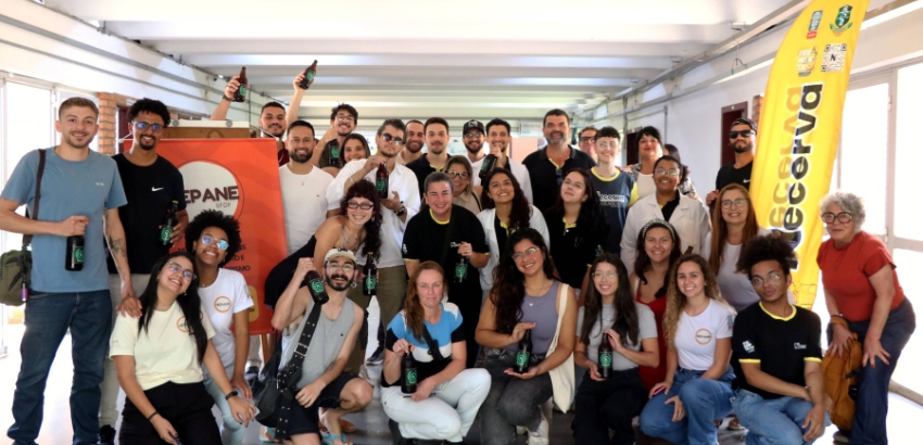 Grupo de participantes posa sorrindo em um corredor após a oficina de harmonização de cervejas com produtos de panificação, realizada pelo Necerva em parceria com o Nepane. A maioria segura garrafas de cerveja e banners dos projetos aparecem ao fundo