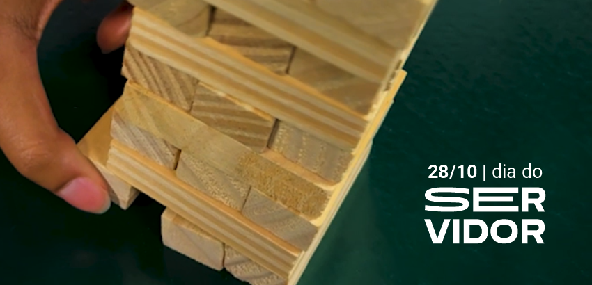 Mão remove peça de torre de blocos de madeira, simbolizando equilíbrio e sustentação. Ao lado, texto “28/10 | dia do servidor” destaca a homenagem ao Dia do Servidor Público
