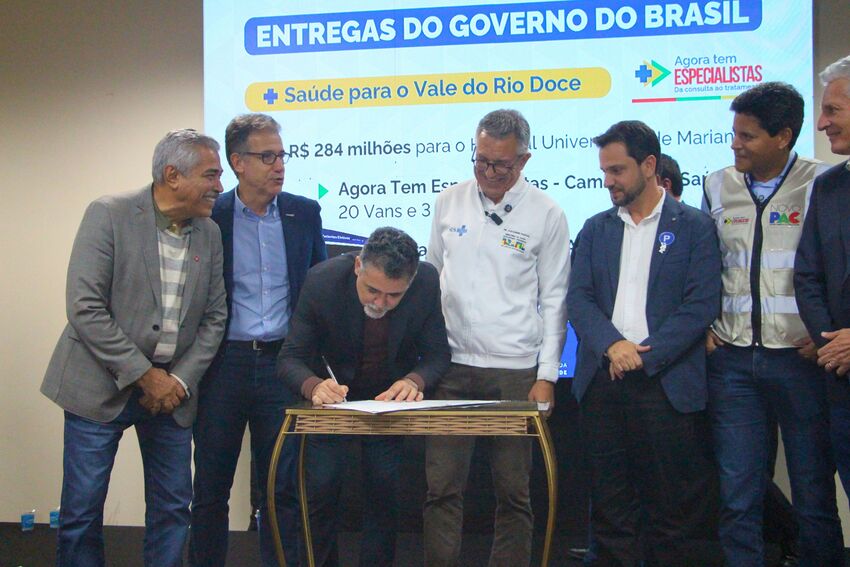 Fotografia de uma cerimônia institucional realizada em auditório. No centro da imagem, um homem inclinado sobre uma pequena mesa assina um documento com uma caneta. Ao seu redor, sete homens observam a assinatura, alguns sorrindo e conversando entre si. O