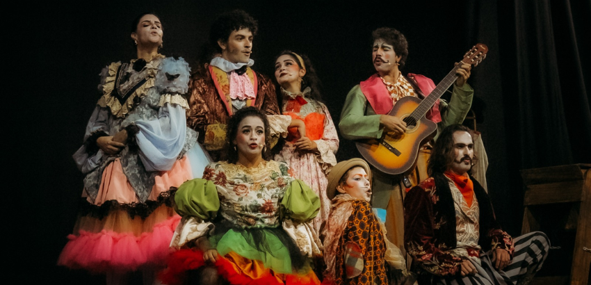 Foto de apresentação teatral com sete atores caracterizados com figurinos de época, em um palco escuro. Ao centro, uma atriz com vestido verde; à direita, um ator toca violão.