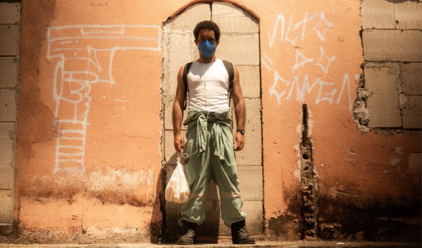 Homem em pé diante de parede alaranjada desgastada, com pichações - uma delas desenha uma arma e outra traz palavras em branco. Ele usa máscara azul, camiseta branca e calça verde amarrada na cintura, segurando uma sacola plástica sob luz intensa do sol.