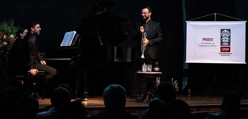 Apresentação musical com dois músicos sentados ao piano à esquerda e um músico em pé com saxofone à direita