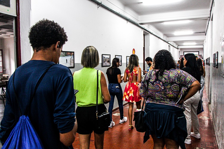 Pessoas observando a exposição fotográfica realizada no Instituto de Ciências Sociais (ICSA) em 2024. A imagem mostra, um corredor do prédio Padre Avelar, onde estão expostas nas paredes quadros com as fotografias dos estudantes. 