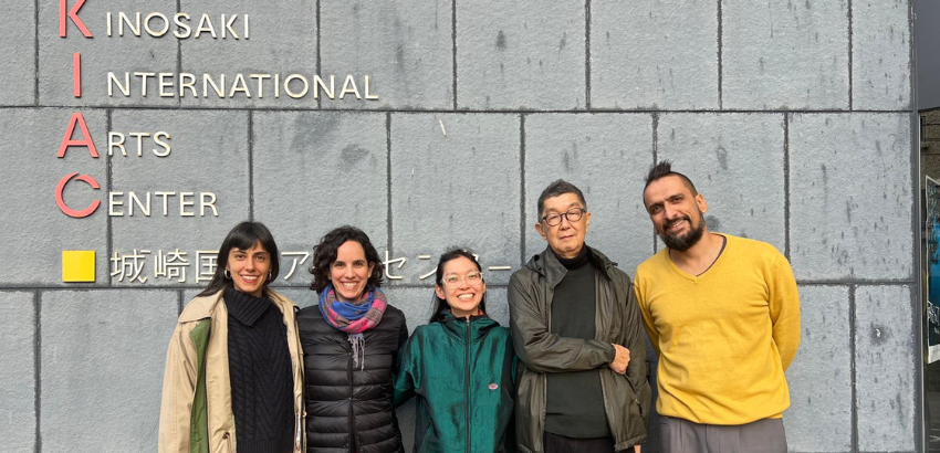 Cinco pessoas estão em pé sorrindo em frente a uma parede cinza com o letreiro “Kinosaki International Arts Center”. Elas usam roupas de frio e parecem posar para uma foto de grupo.