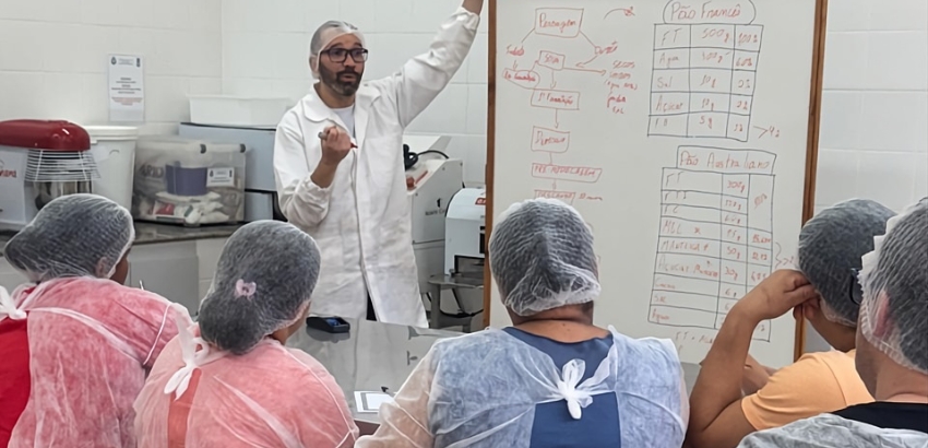 Instrutor de jaleco branco explica conteúdo em um quadro branco para um grupo de alunos usando toucas e aventais descartáveis em uma sala de panificação, com equipamentos de cozinha industrial ao fundo.