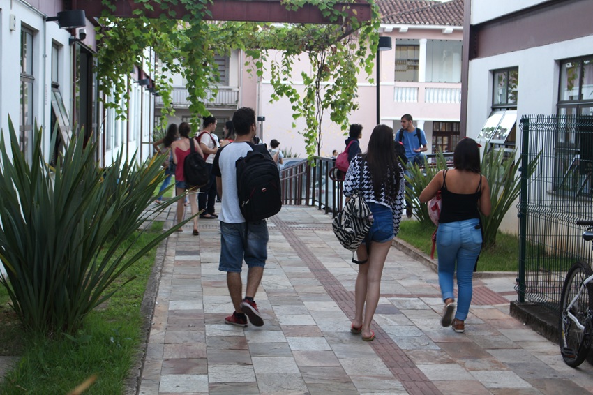 Estudantes caminham por corredor arborizado entre prédios do Instituto de Ciências Sociais Aplicadas da UFOP. Alguns carregam mochilas e bolsas, criando um ambiente de convivência acadêmica no campus.