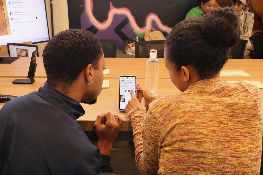 Durante uma atividade prática em um workshop, um bolsista do projeto auxilia uma participante com o uso de uma rede social em seu celular. Eles estão sentados lado a lado em uma mesa de reunião, concentrados na tela do aparelho.