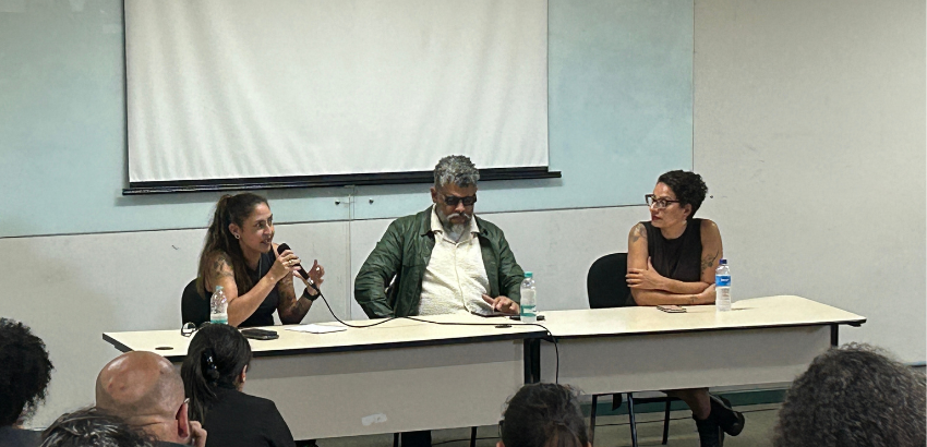 Mesa com três participantes em um debate, com uma mulher ao microfone e público à frente.