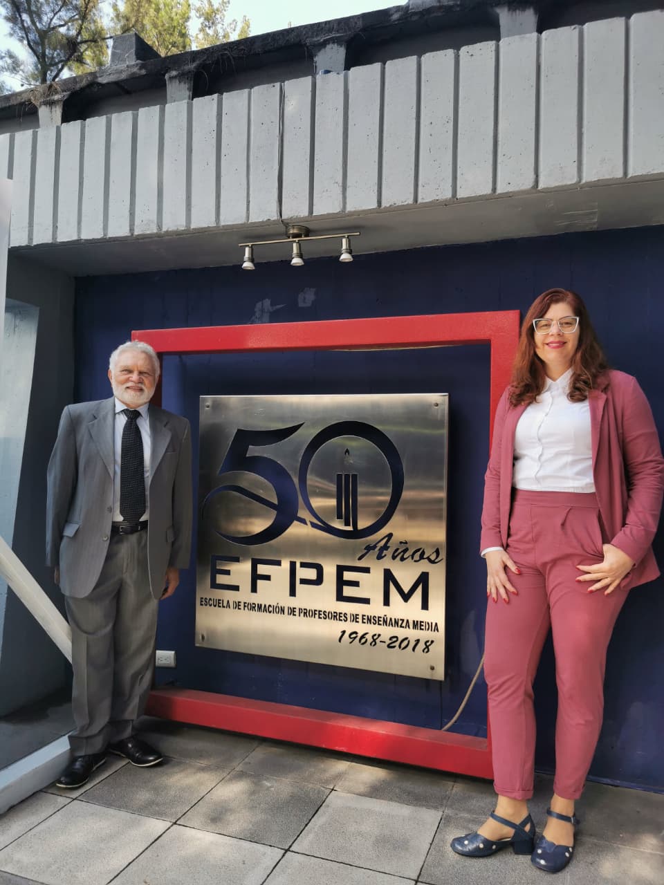 Imagem mostra os professores José Rubens Lima Jardilino e Paula Cristina Cardoso Mendonça, em visita à Guatemala. O professor usa terno cinza , gravata escura. A professora usa rosa e camisa social branca, eles estão em frente a placa de 50 anos da EFPEM.