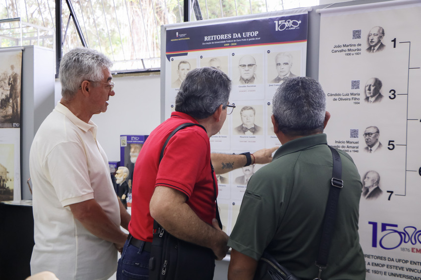 Três homens observam um painel com fotografias e informações sobre os reitores da UFOP.