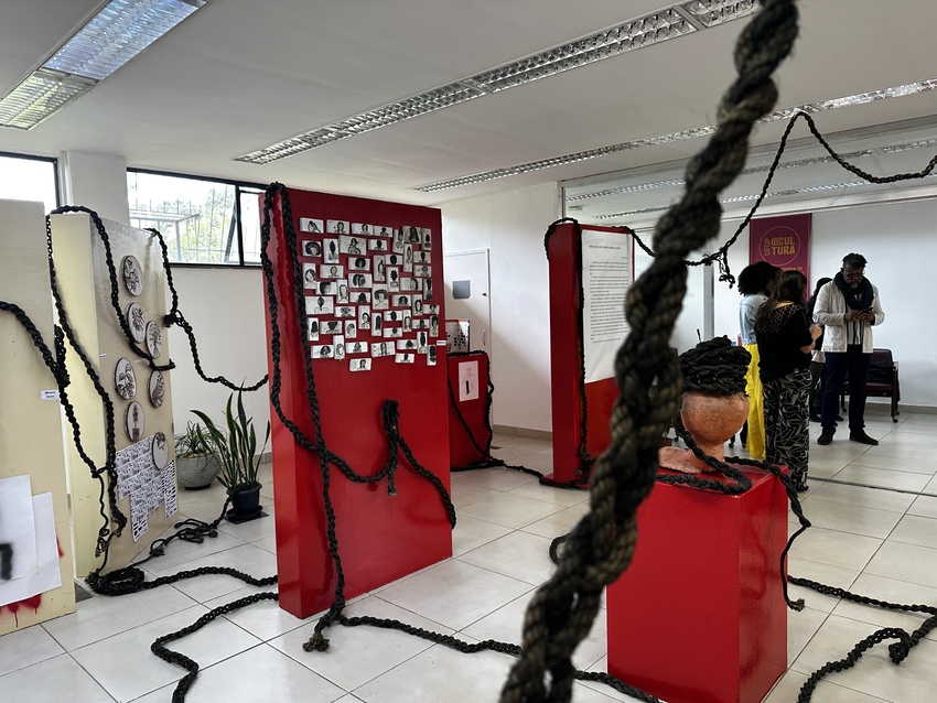 Sala expositiva com painéis vermelhos e brancos ocupando o espaço. Cordas grossas e trançadas se espalham pelo chão, sobem pelas estruturas e atravessam o ambiente, criando uma instalação artística imersiva. Em um dos painéis há dezenas de fotografias em 