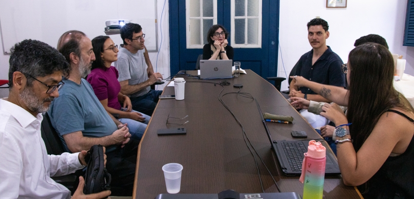 Reunião em uma sala com mesa comprida: oito pessoas sentadas dos dois lados, conversando e trabalhando com notebooks, celulares e anotações