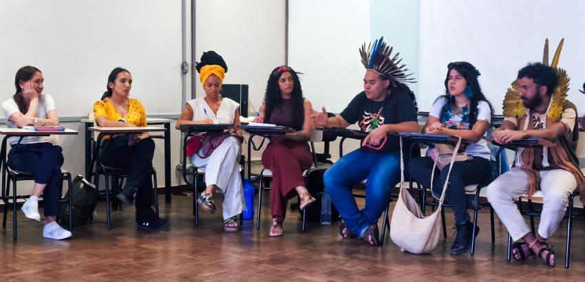 Grupo de pessoas sentadas em círculo em uma sala de aula, participando de uma roda de conversa. Alguns usam cocares e adornos indígenas, enquanto outros anotam ou ouvem atentamente.
