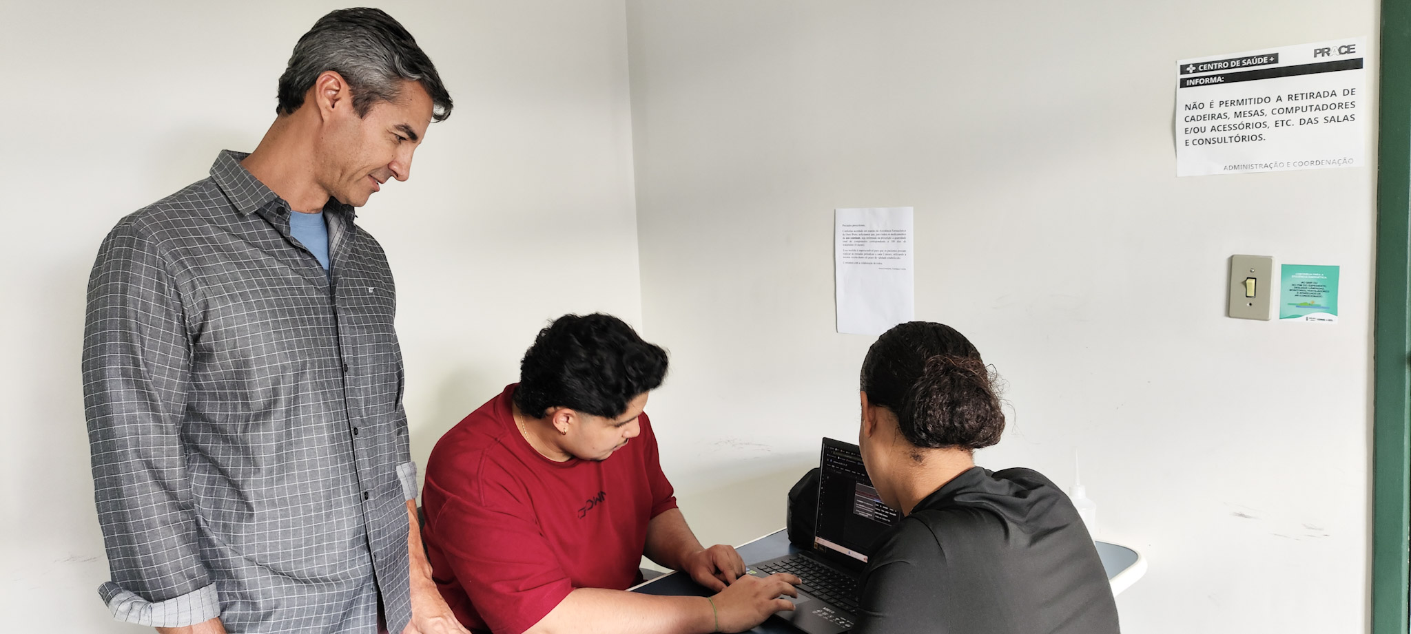 Homem em pé observa duas pessoas sentadas trabalhando em um notebook dentro de uma sala de atendimento de uma unidade de saúde. A pessoa de camiseta vermelha digita no computador enquanto a profissional à frente acompanha a tela. Ao fundo, há paredes clar
