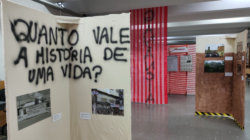 Exposição montada em um espaço interno, com painéis de tecido e fotografias que retratam famílias e locais atingidos. Em destaque, uma parede traz a frase “QUANTO VALE A HISTÓRIA DE UMA VIDA?” escrita em tinta preta. Ao fundo, há tecidos listrados em verm