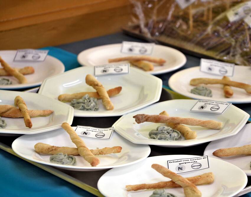 Pratos brancos alinhados sobre uma mesa exibem grissinis artesanais acompanhados de um creme de gorgonzola. Pequenas etiquetas identificam o alimento. Cada prato está com duas unidades de grissini e uma porção de creme ao lado