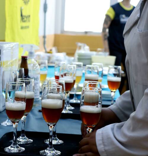 Pessoa de jaleco segura uma bandeja com taças de cerveja âmbar com espuma, enquanto várias outras taças iguais estão dispostas sobre uma mesa ao fundo