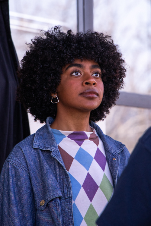 Mulher negra com cabelo cacheado e camiseta colorida olha para cima com expressão atenta, em um ambiente com luz natural. Está usando brincos de argola e uma jaqueta jeans.