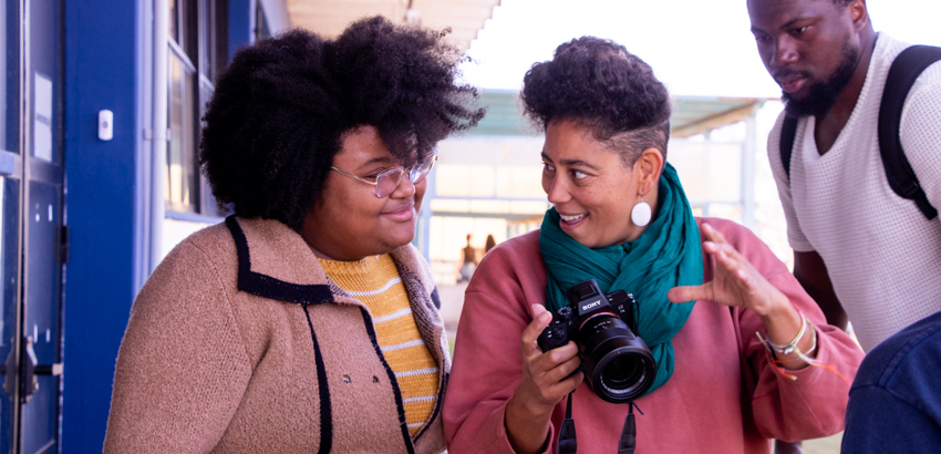 Três pessoas conversam ao ar livre, uma delas segura uma câmera e gesticula explicando algo. As outras duas observam atentamente, sorrindo, em um momento de troca de ideias.