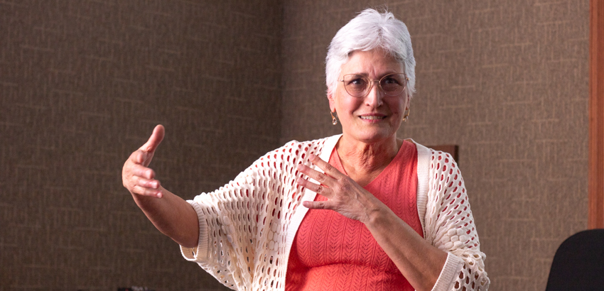 Pessoa de cabelos curtos e brancos, usando óculos, blusa coral e casaquinho branco. Em ambiente interno, gesticula com uma mão estendida e a outra sobre o peito, transmitindo expressividade e envolvimento na fala.