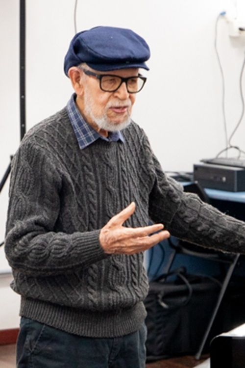O compositor Rufo Herrera, de boina azul e suéter de tricô cinza, fala com entusiasmo durante a homenagem no curso de Música da UFOP, gesticulando com as mãos enquanto compartilha memórias e reflexões de sua trajetória.